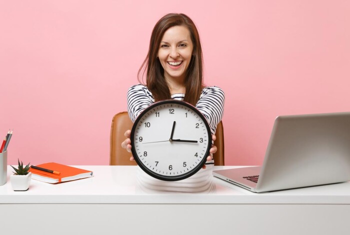 pq-smiling-woman-holding-round-alarm-clock-while-sit-working-project-white-desk-with-contemporary-pc-laptop