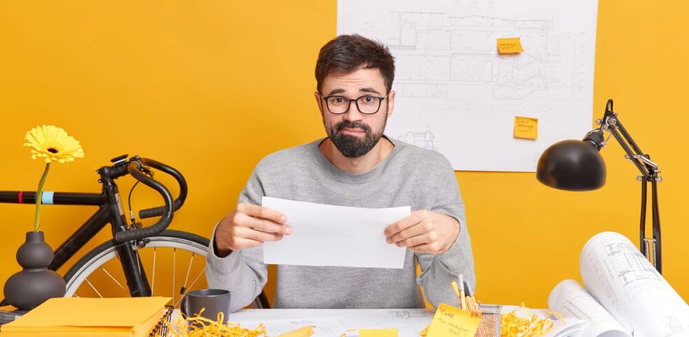 puzzled-bearded-skilled-male-illustrator-holds-paper-has-troubles-with-future-project-poses-coworking-space-has-mess-desk-man-freelance-architect-prepares-new-house-development-drawings-pq